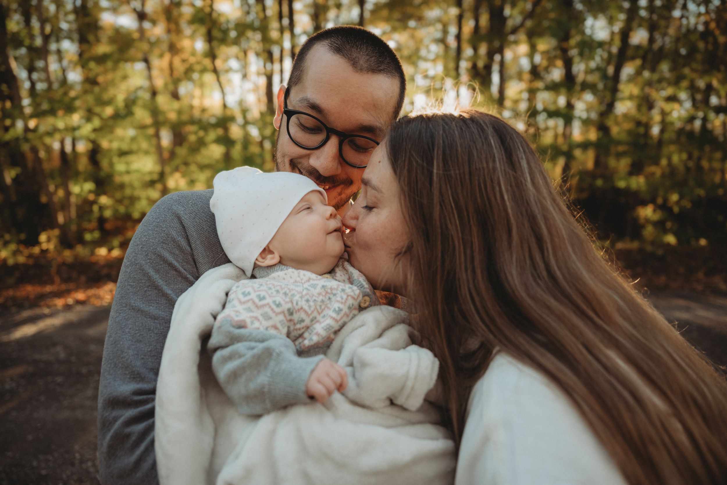 A tender family moment as parents kiss their baby's cheeks in an autumn forest setting.