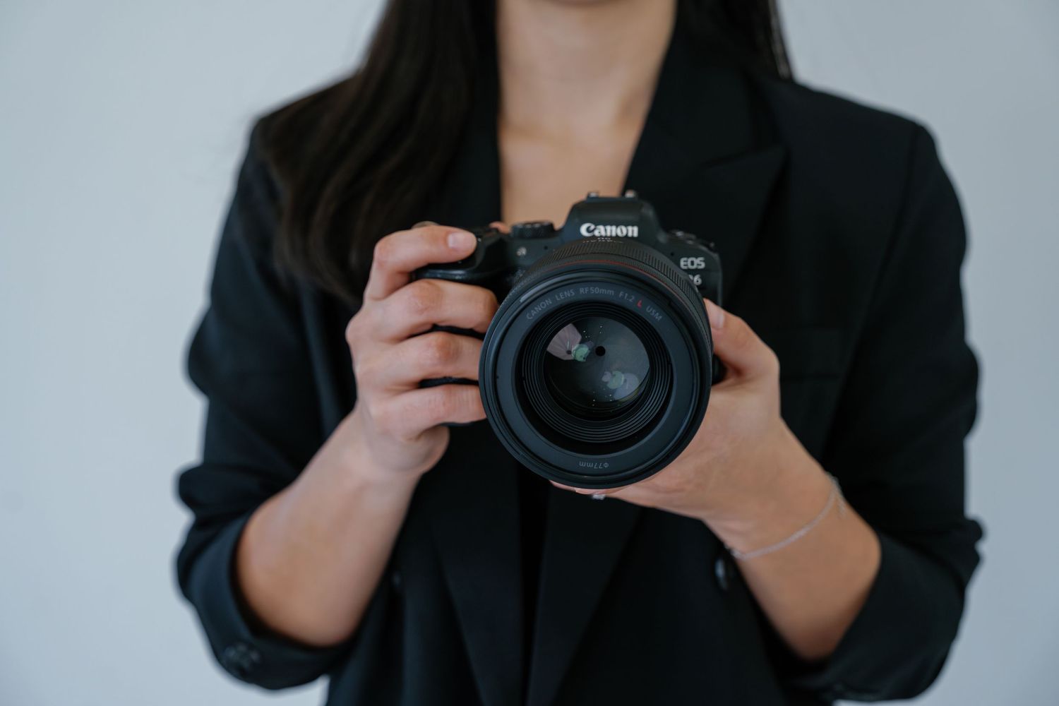 Person in black blazer holding a Canon DSLR camera with professional lens against white background.