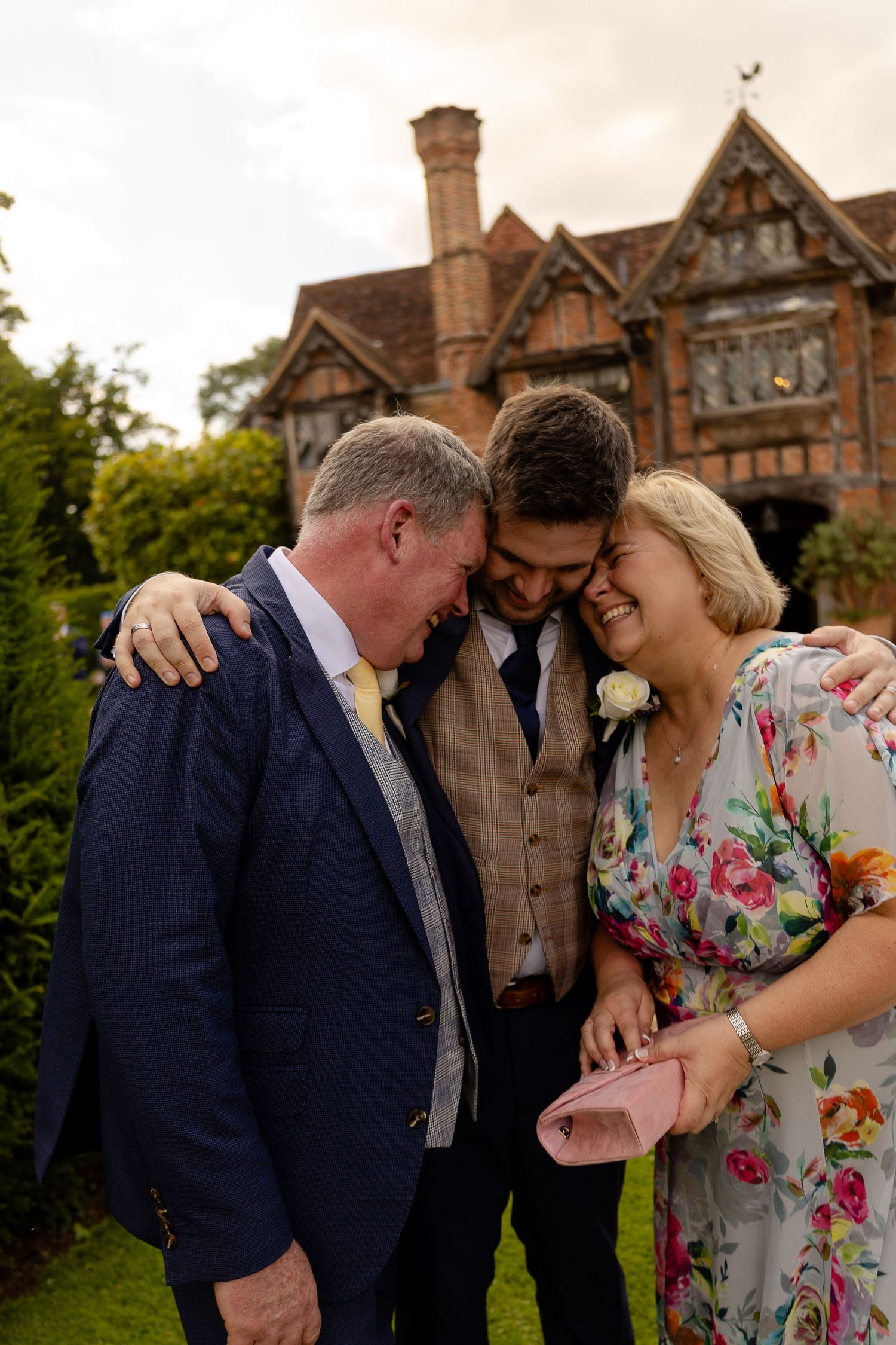 A joyful shared moment among three loved ones in front of a Tudor-style mansion on a summer evening.