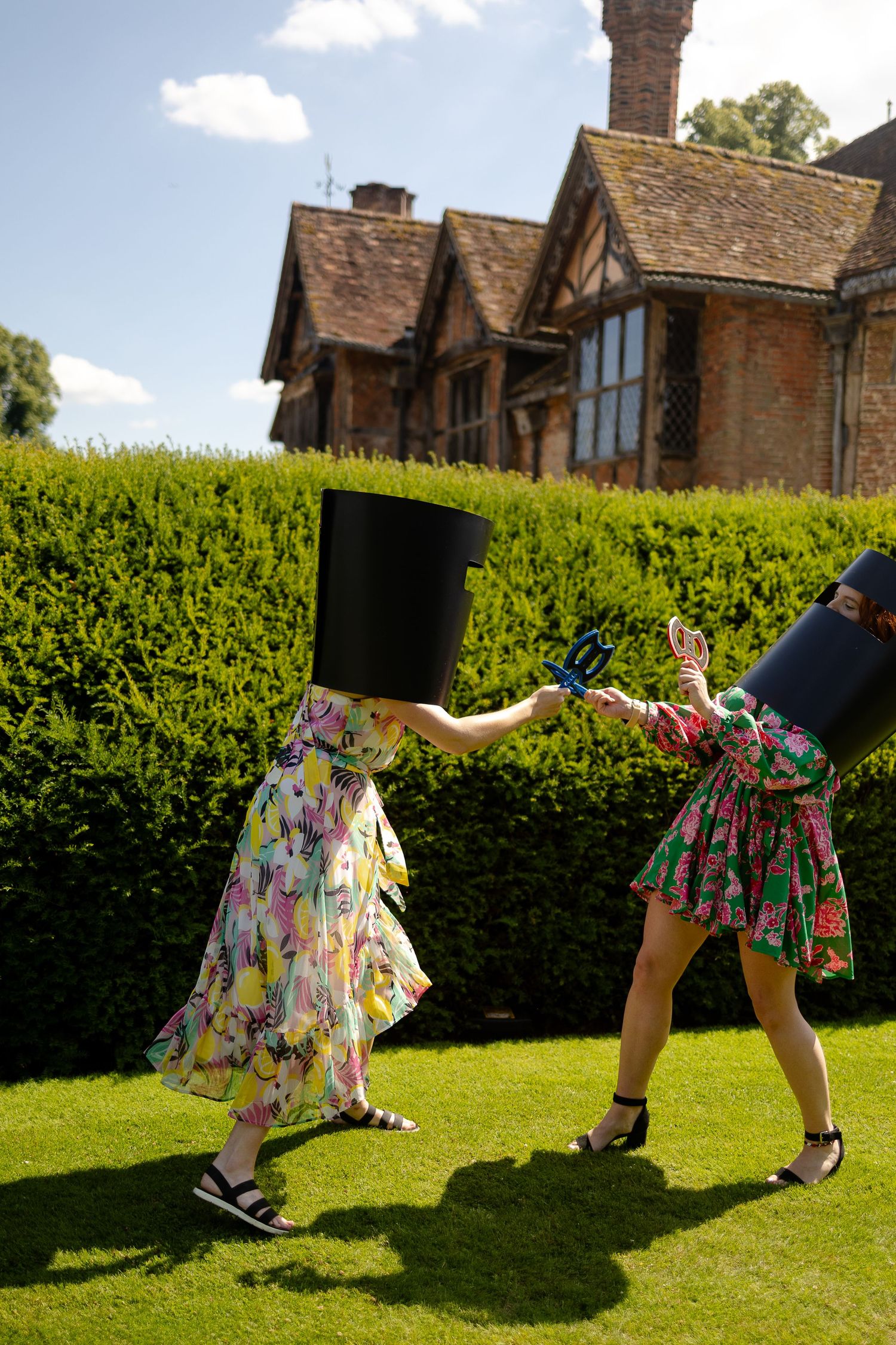 Two people in floral dresses dance playfully on a lawn with a tall top hat in front of a Tudor-style house.