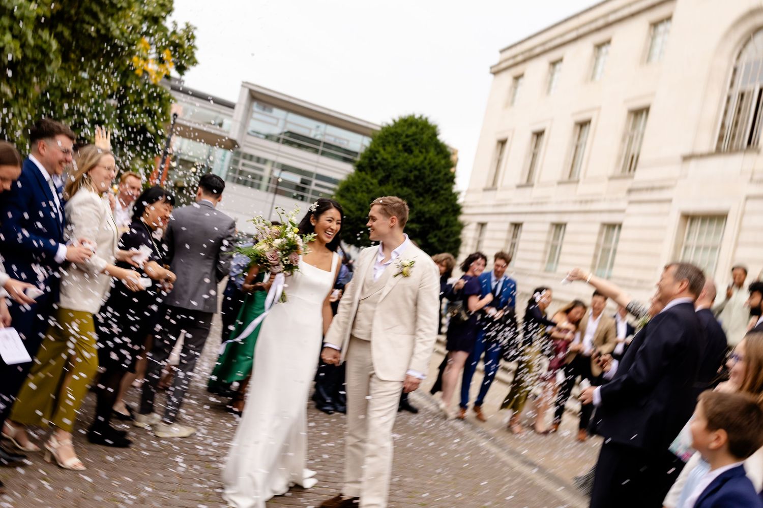 A couple stands surrounded by wedding guests throwing confetti on a cobblestone courtyard outside an elegant building.