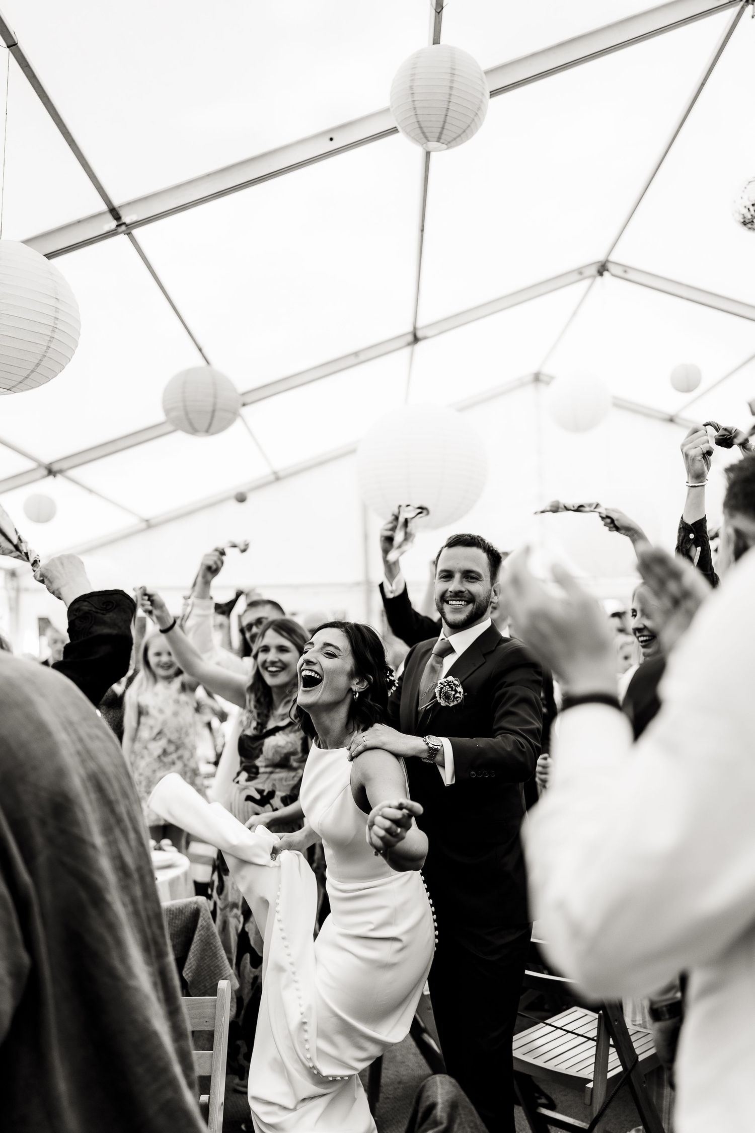 Wedding celebration under a tent with paper lanterns as guests dance and cheer during a lively reception moment.