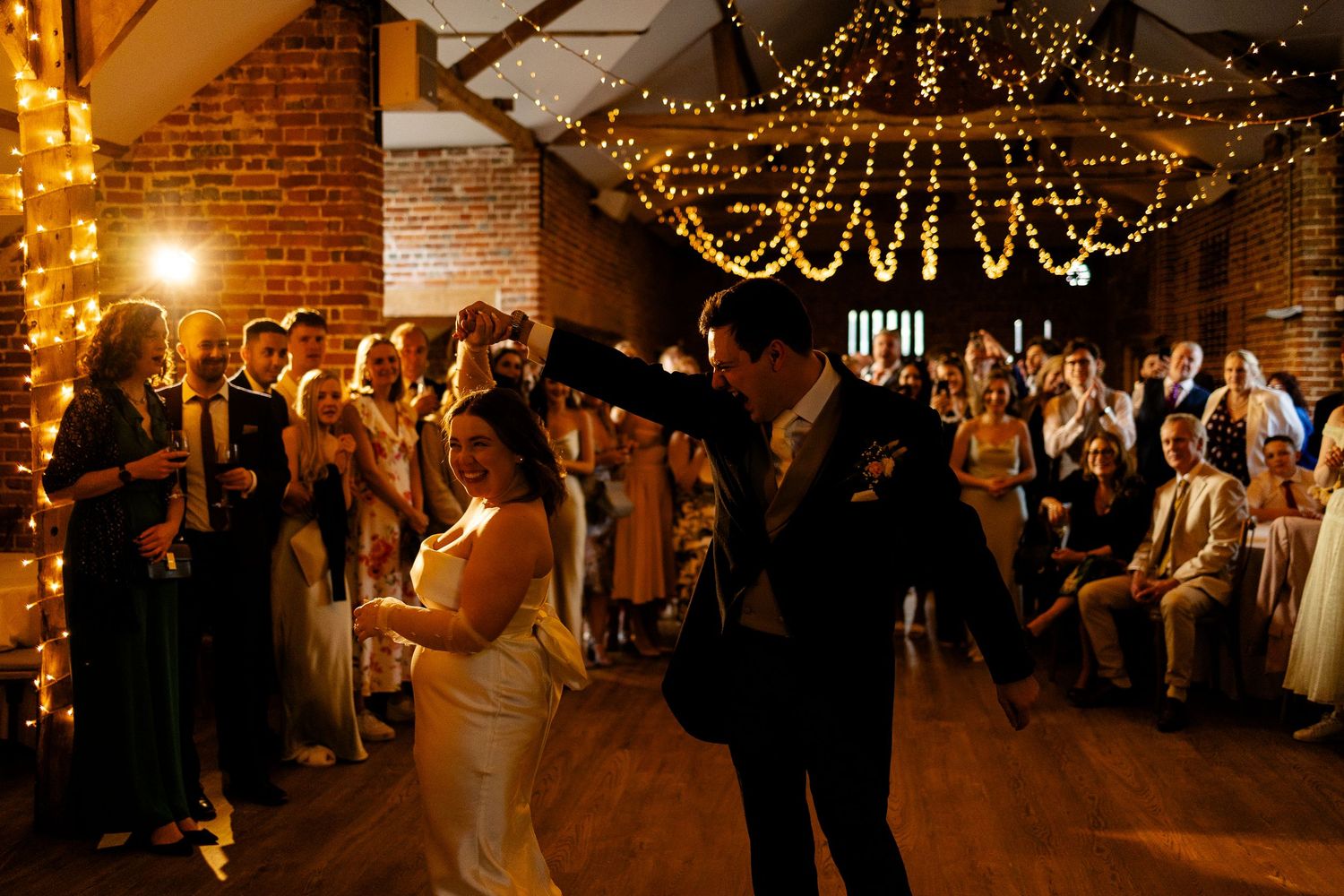 Guests dance beneath twinkling string lights at a rustic indoor wedding reception with exposed brick walls.