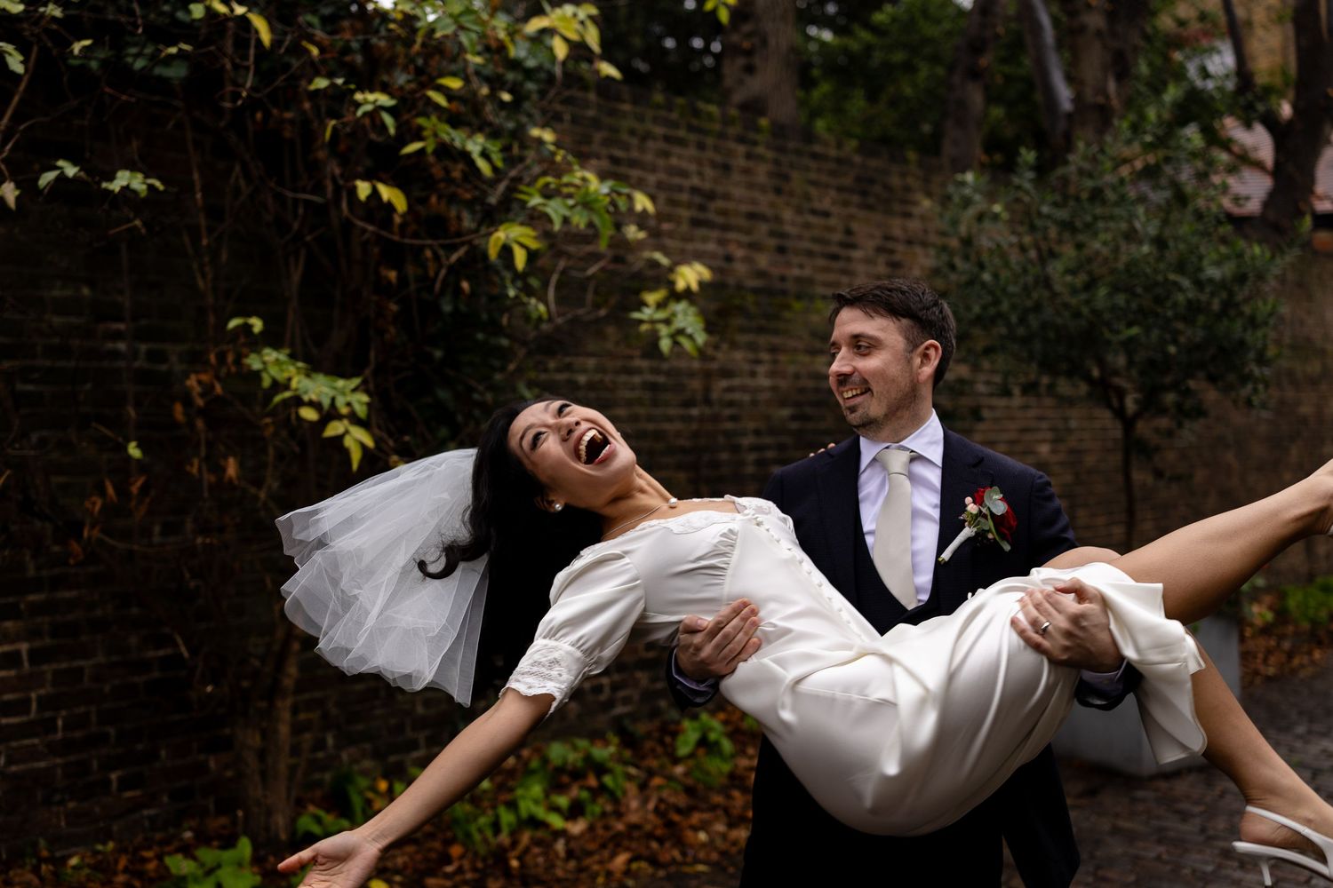 A joyful couple poses outdoors during their wedding day, with the bride leaning back dramatically in the groom's arms.