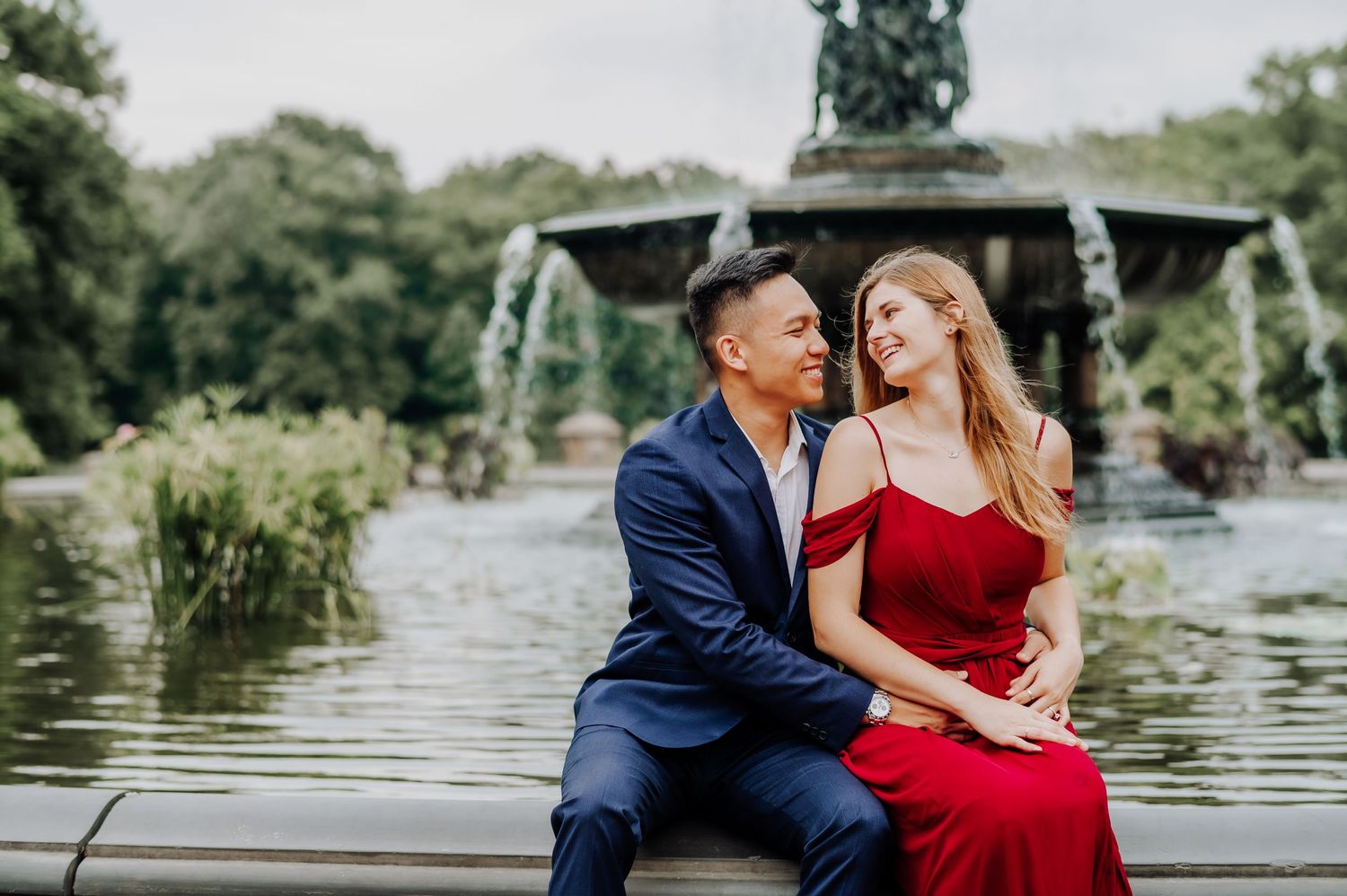 Romantic couple on dock with woman in red dress and man in navy suit against pond backdrop.