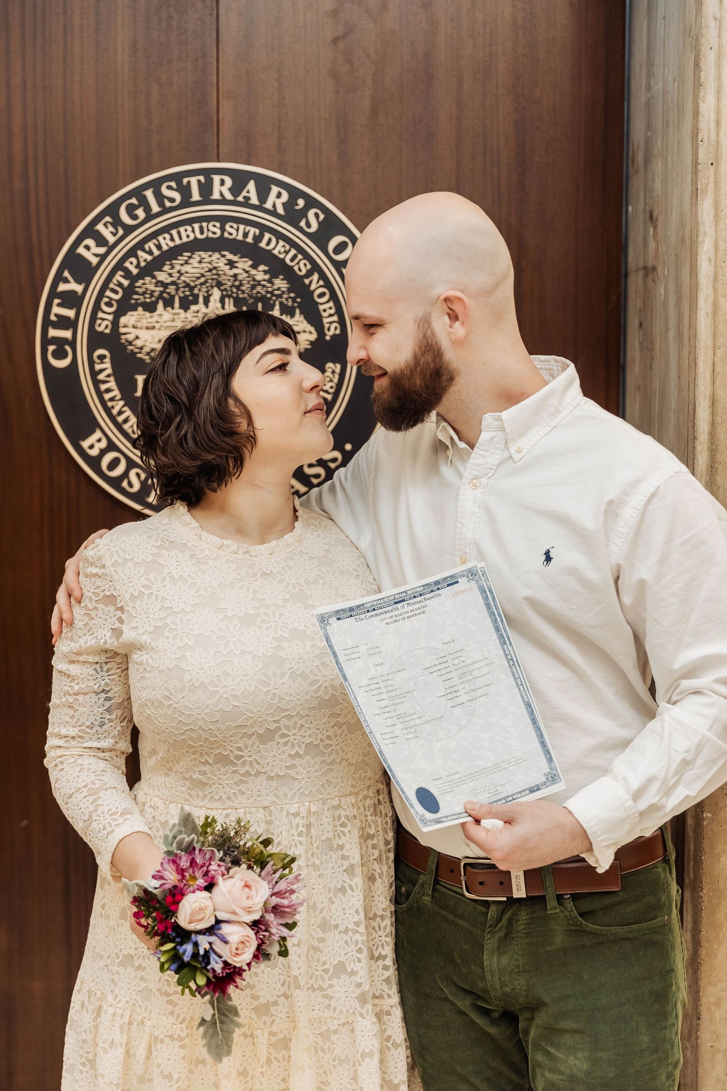 A couple stands before a city registrar's seal holding their marriage license and a floral bouquet.