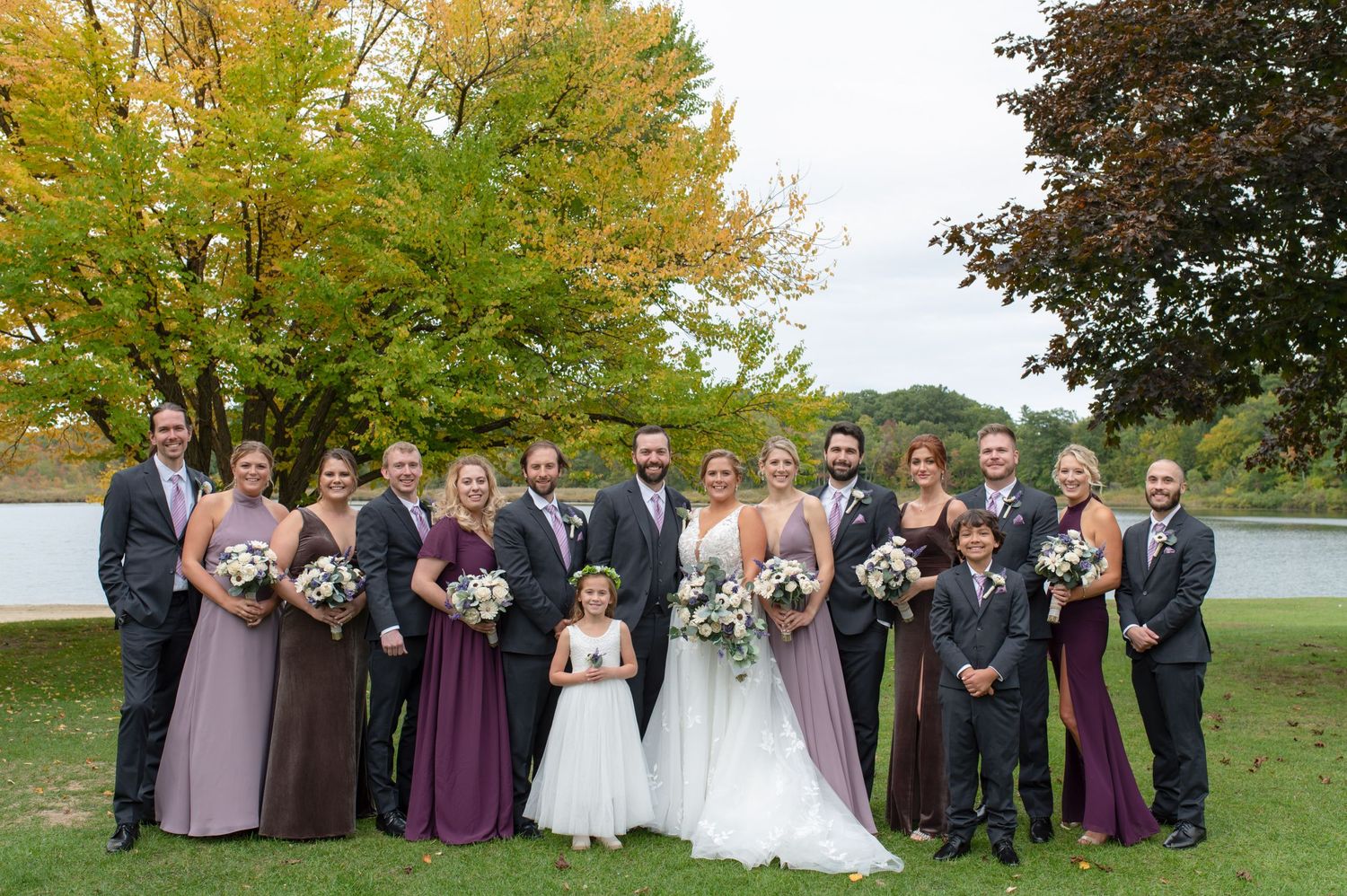 Wedding party poses outdoors in coordinated purple and gray attire against fall foliage.