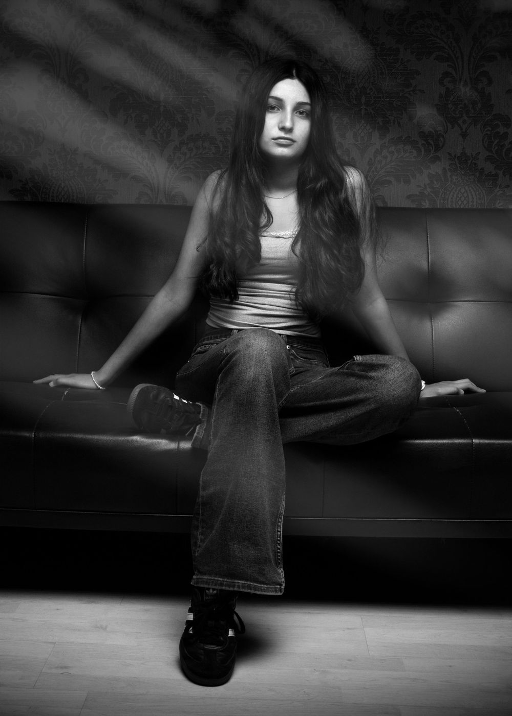 A dramatic black and white portrait of a person in flared jeans and a tank top seated on a leather couch.