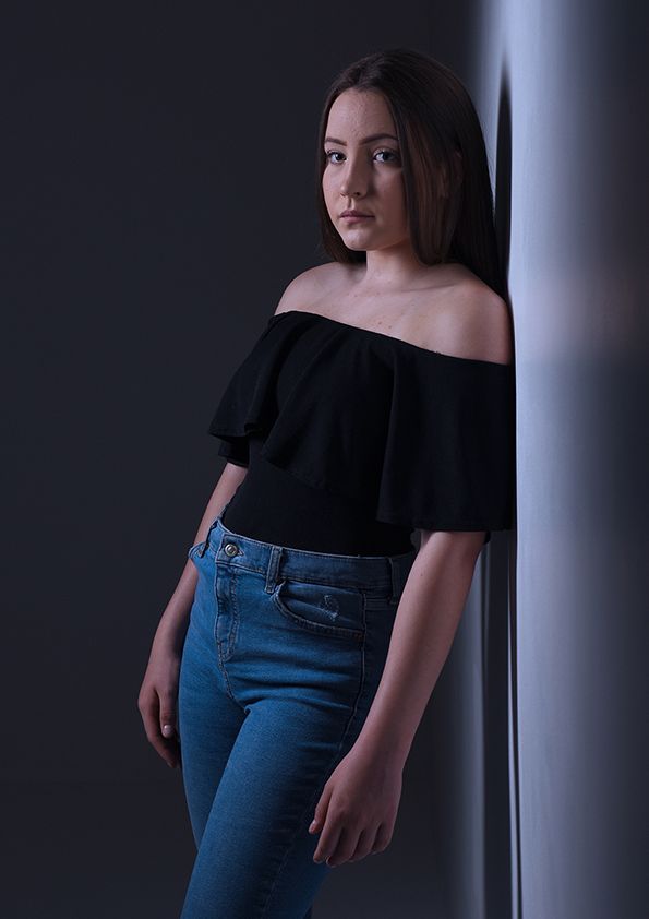Moody portrait against dark wall wearing off-shoulder top and blue jeans.