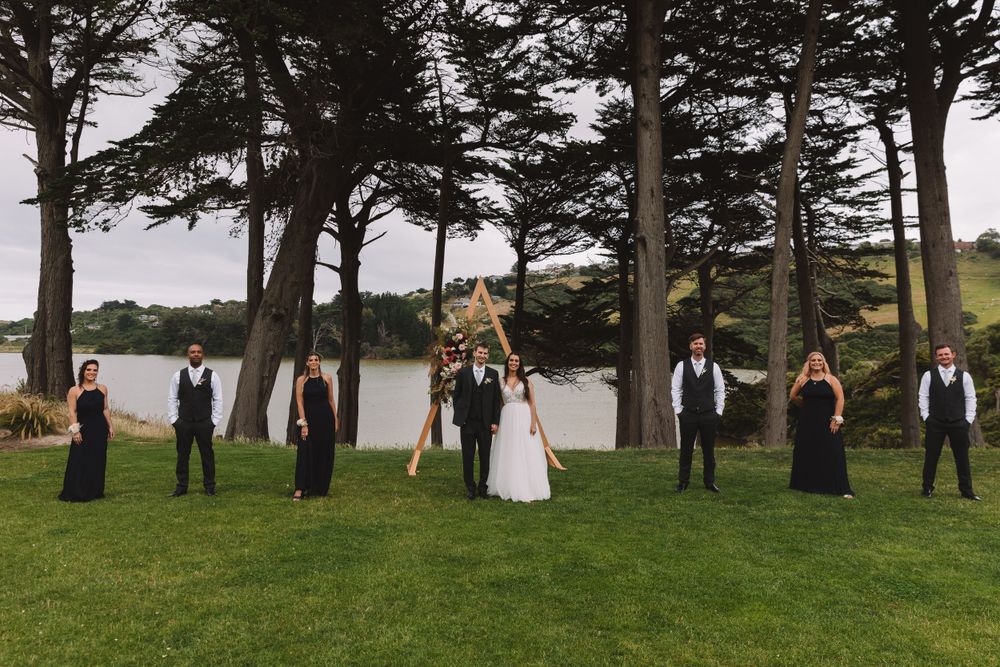 Wedding party poses formally under tall pine trees beside a lake with a wooden triangular arch.