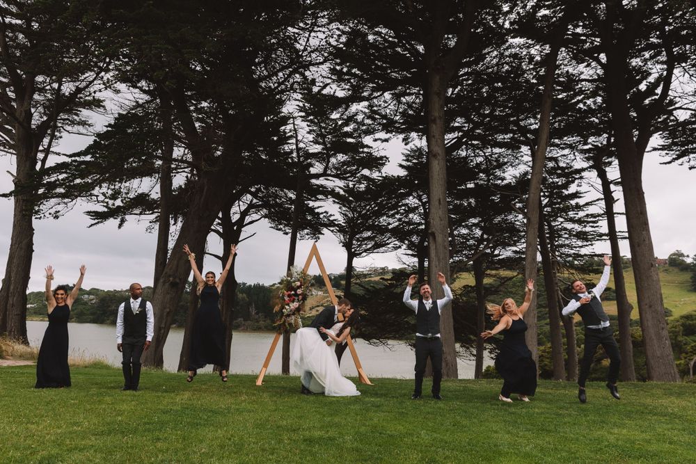 Wedding party celebrates with arms raised under pine trees by the water.