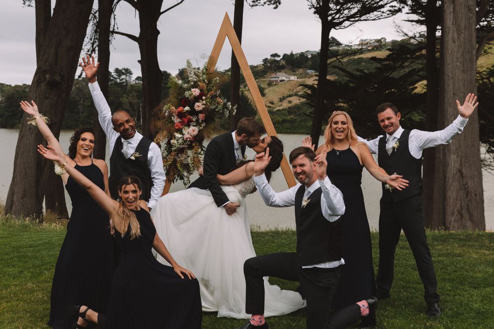Wedding party poses playfully with triangular arch in background near waterfront.