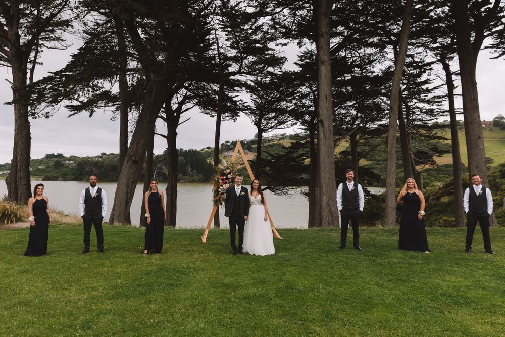 Wedding group stands symmetrically on grass with triangular arch and pine trees framing the scene.
