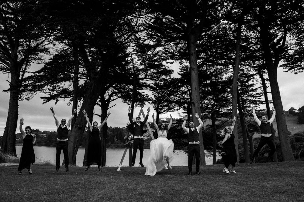 Black and white photo of wedding party jumping with joy outdoors by the water.