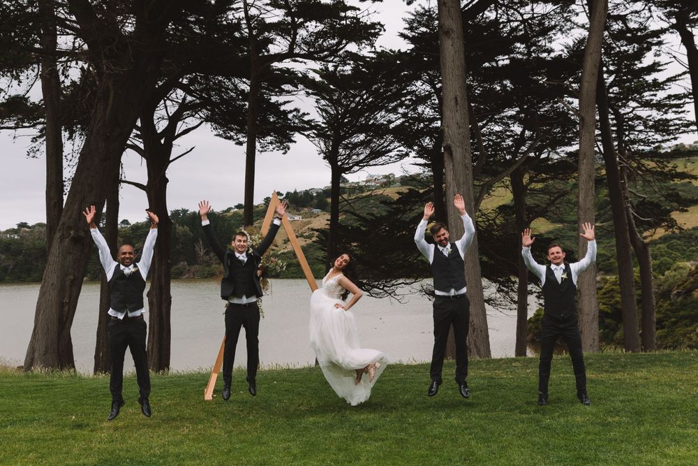 Wedding party celebrates joyfully on a grassy lawn with tall pine trees and water in the background.