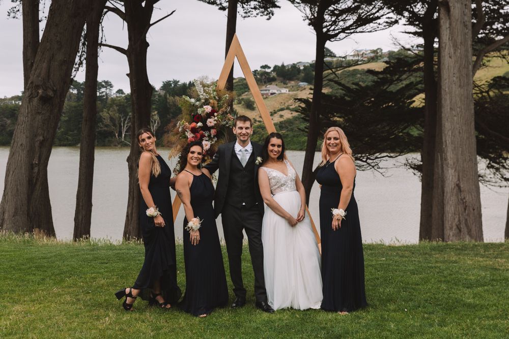 Wedding party poses for formal group photo near a lake surrounded by evergreen trees.