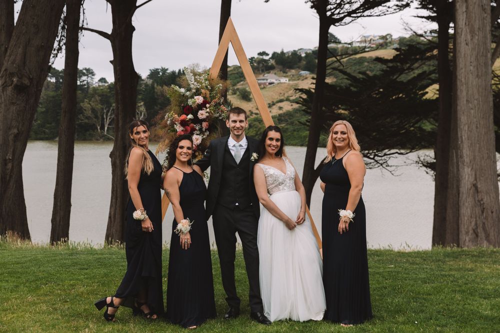 Wedding group poses with wooden triangle arch decorated with flowers by lakeside.