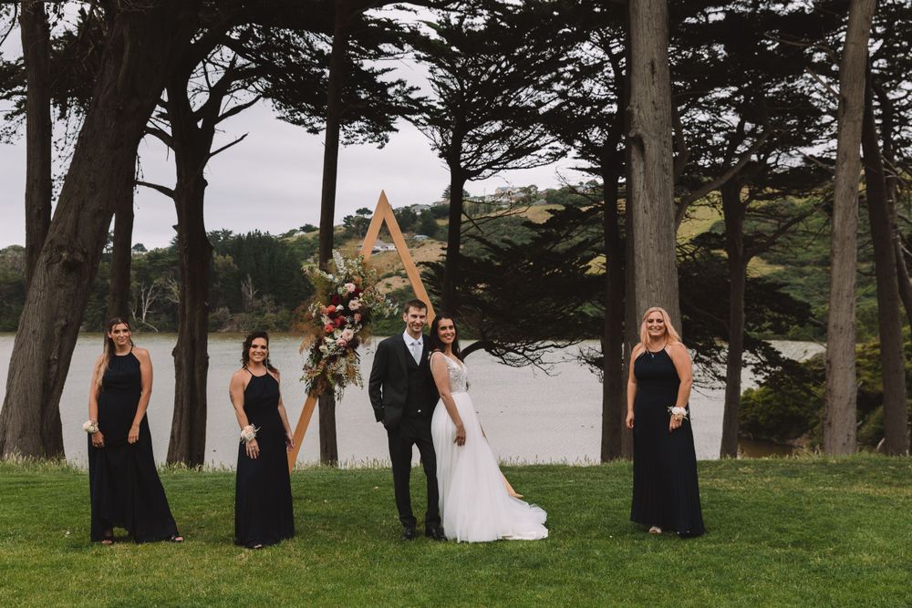 Wedding ceremony setup with wooden triangle arch and black-dressed attendants by the lake.