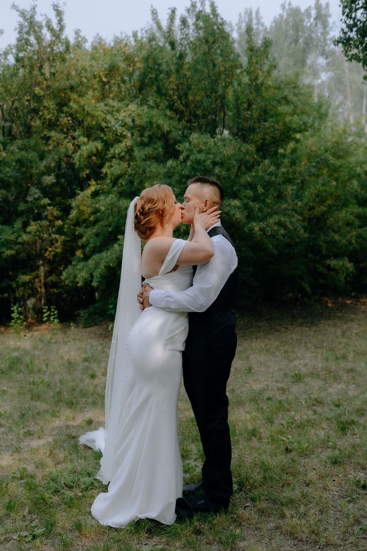 A romantic embrace between partners in wedding attire against a backdrop of green foliage.