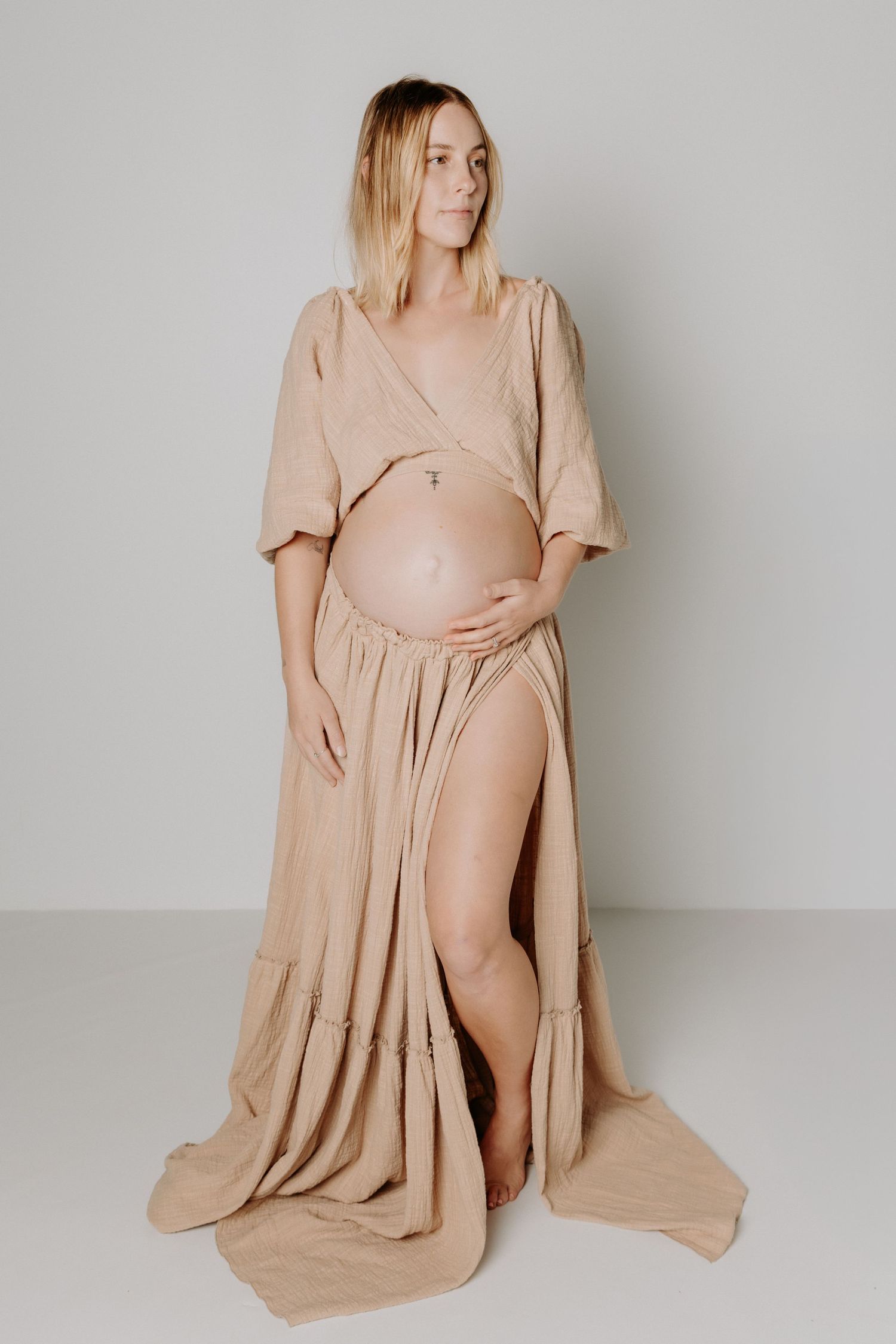 Maternity photoshoot featuring a flowing tan dress against a minimalist gray backdrop.
