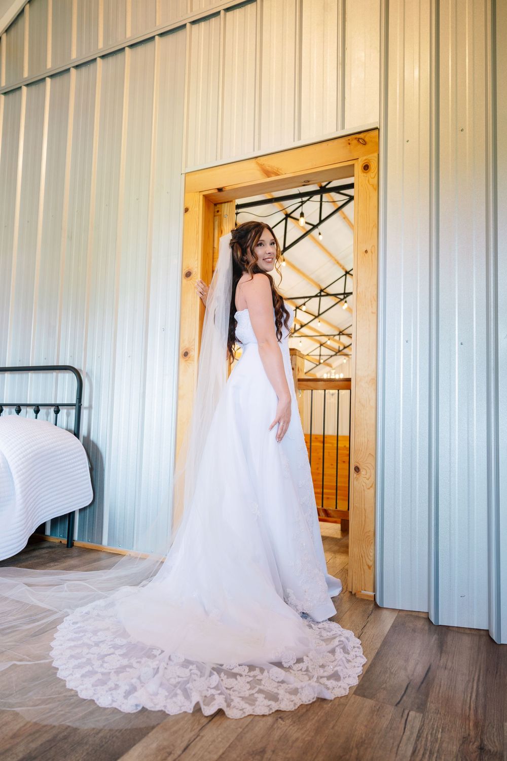 A bride in an elegant white lace wedding gown with a long train stands in a doorway of a rustic venue. Emma b's tree farm