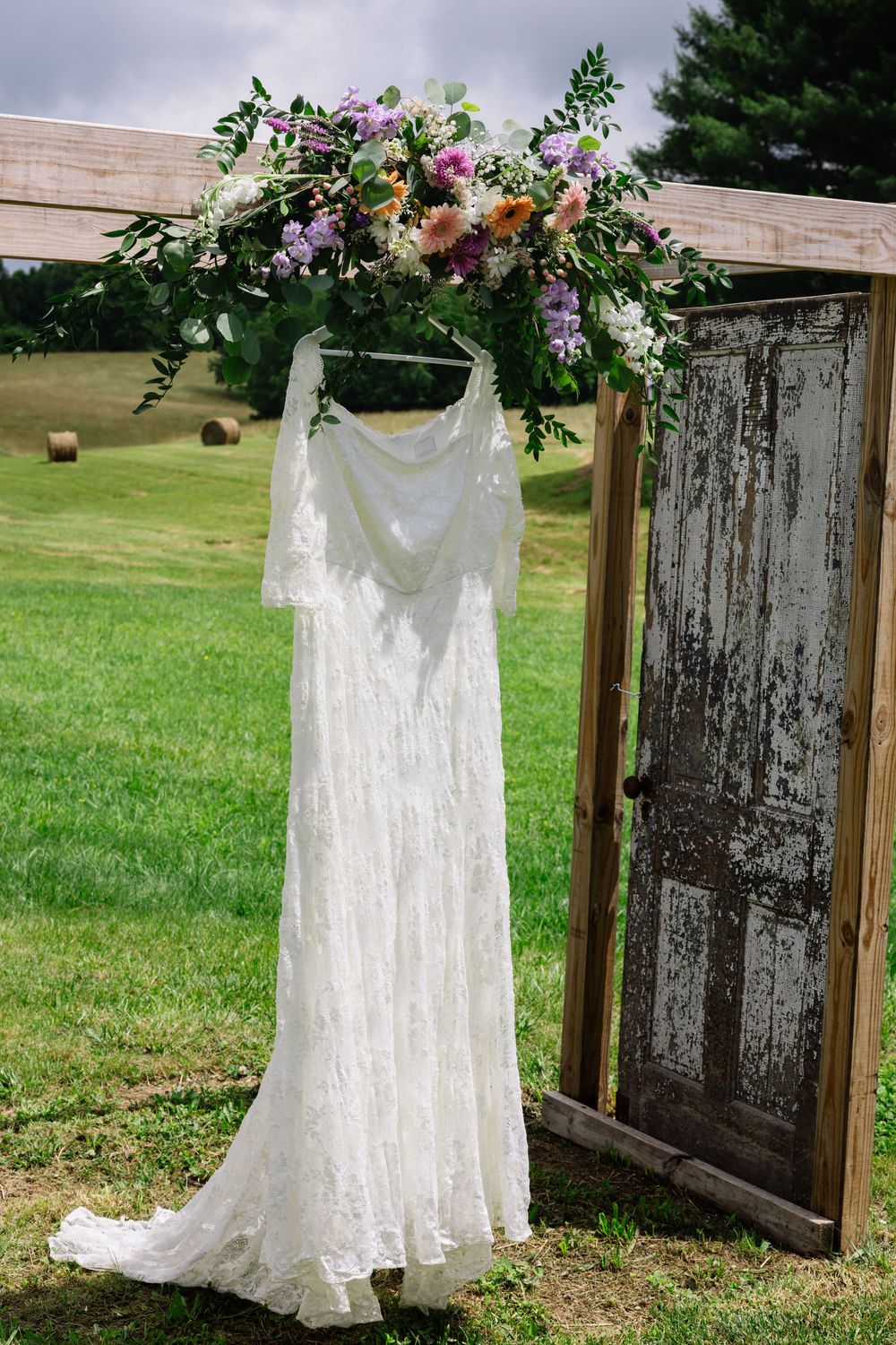 A white lace wedding dress hangs from a wooden arch decorated with pink and purple flowers in an outdoor garden setting. Knoxville.