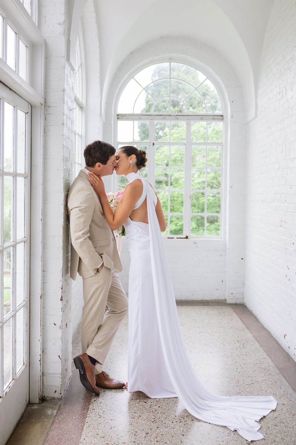 A romantic couple in wedding attire share an intimate moment by a large arched window in a bright white hallway.