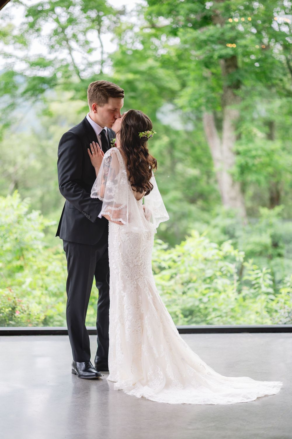 A romantic wedding couple embraces while standing in their formal attire against a lush green forest backdrop.
