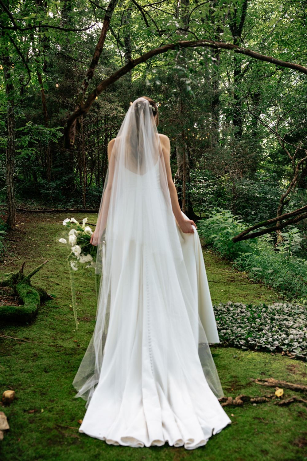 A bride in a flowing white dress and veil stands in a lush forest garden setting surrounded by green foliage and flowers.