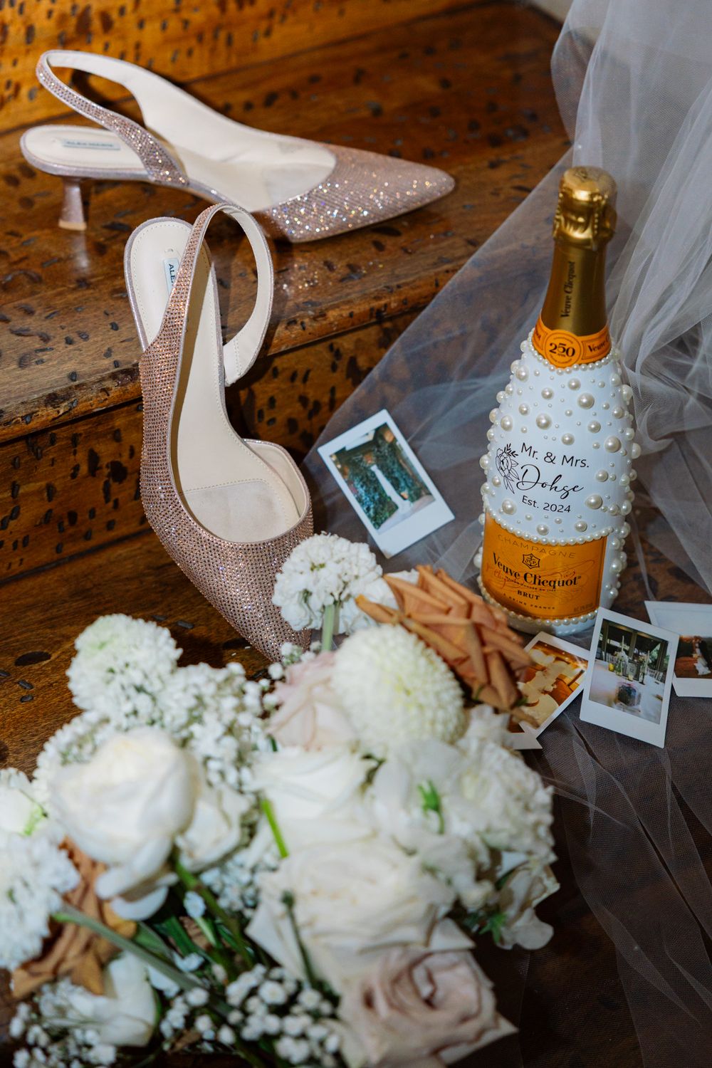 Wedding details including sparkly heels, Veuve Clicquot champagne, pastel roses and Polaroid photos on wooden surface.