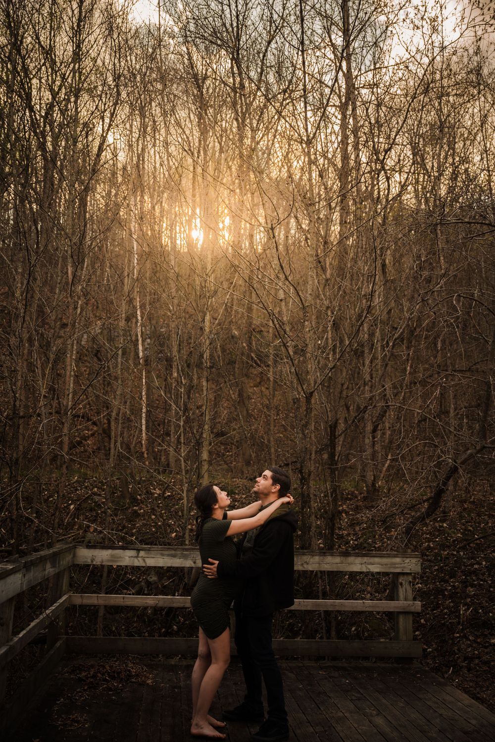 A romantic sunset embrace on a wooden deck surrounded by bare winter trees with golden light filtering through.