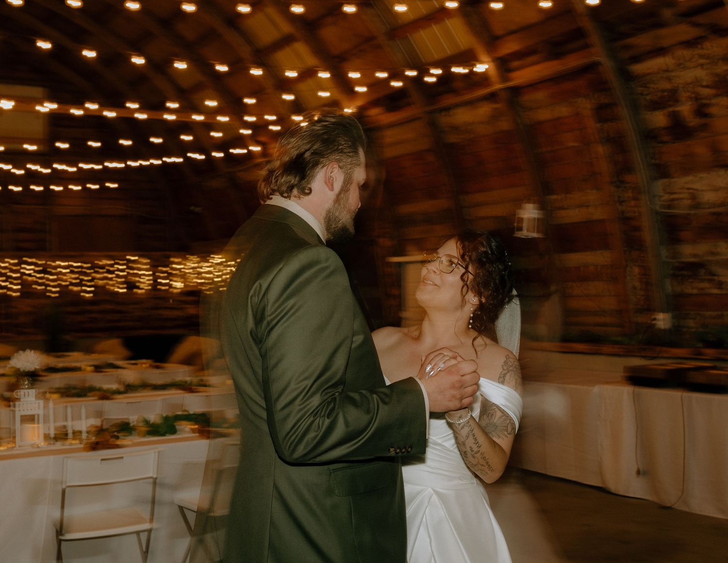 A romantic moment on a dance floor under warm string lights in a rustic barn venue.