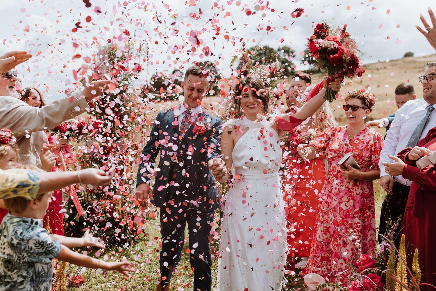 Wedding party celebration with red and white confetti being thrown over a couple during an outdoor ceremony.