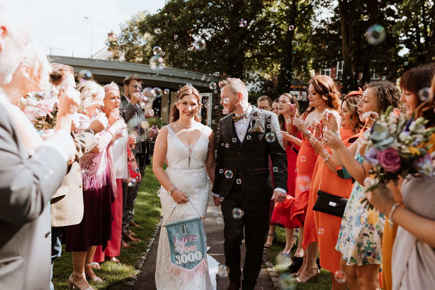Wedding guests throw confetti over newlyweds walking down outdoor aisle during summer garden celebration.