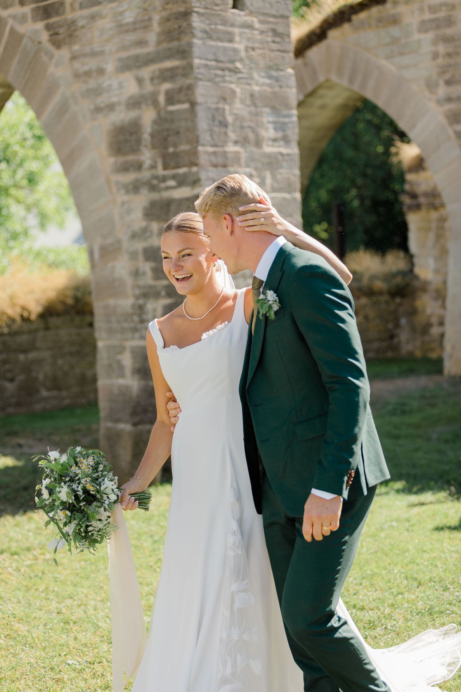 Happy newlyweds share a laugh under stone archways in garden setting.