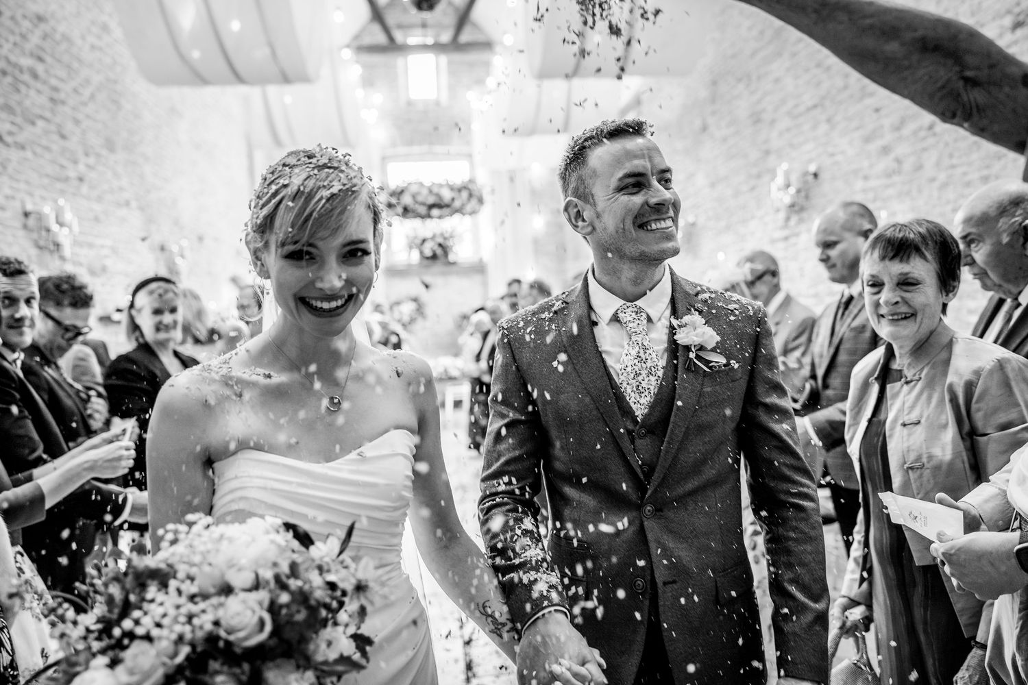 Black and white photo of newlyweds showered with confetti during joyful wedding recessional.