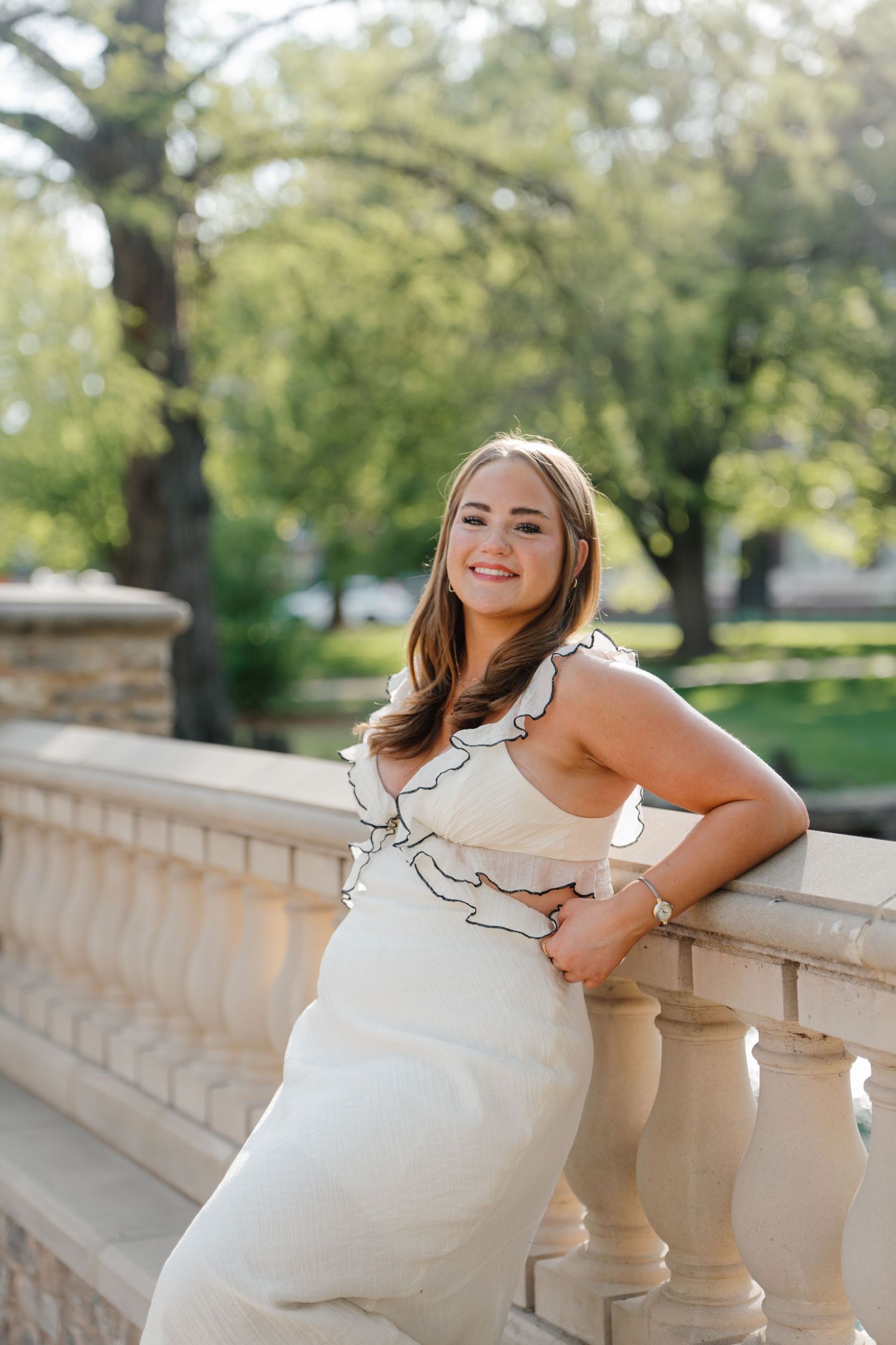 College University Senior in white ruffled dress poses elegantly against stone balustrade, in Stillwater, OK.