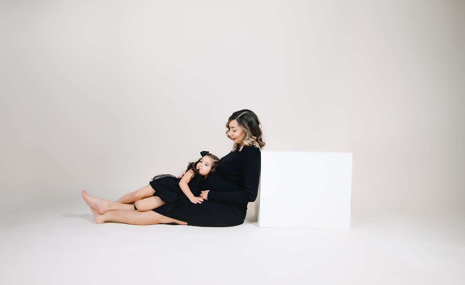 maternity studio photoshoot. simple white backdrop. mother holding her 3 year old daughter while leaning on a posing block. sweet little gir