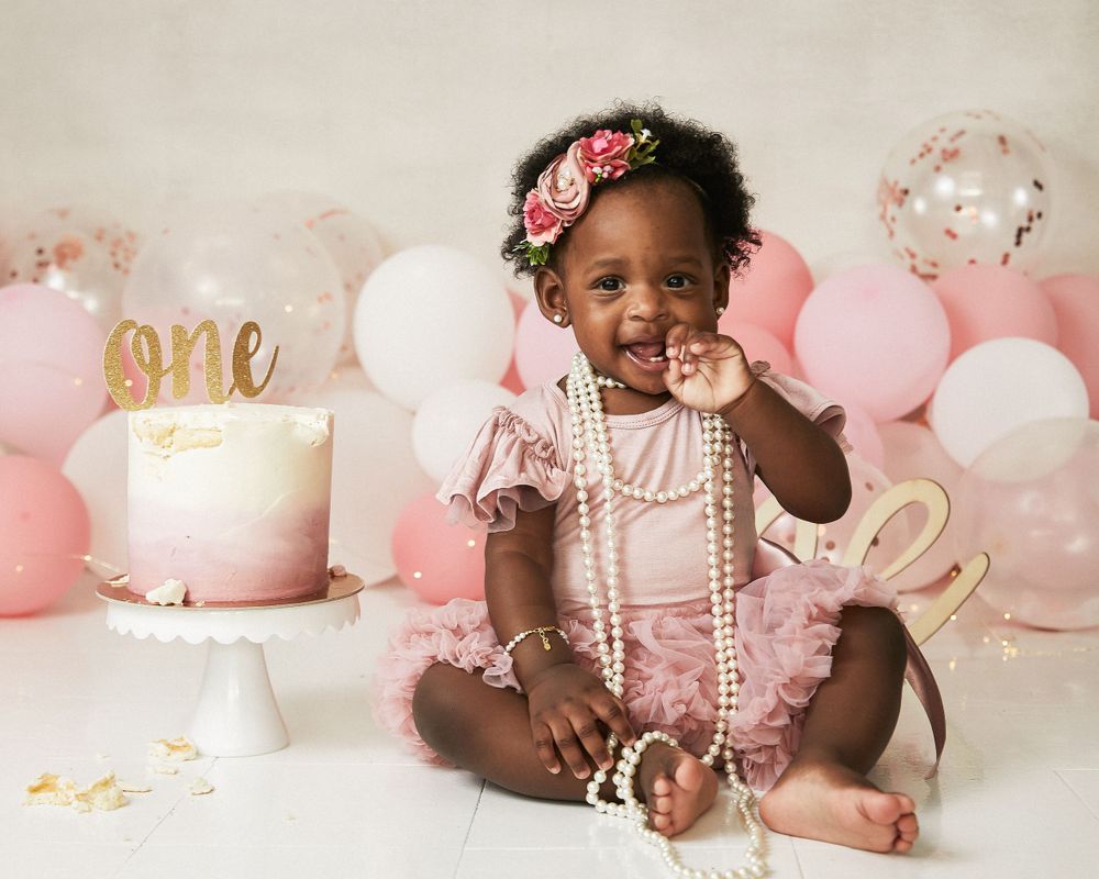 A first birthday celebration with pink balloons, cake, and a toddler in pearls and tulle dress.