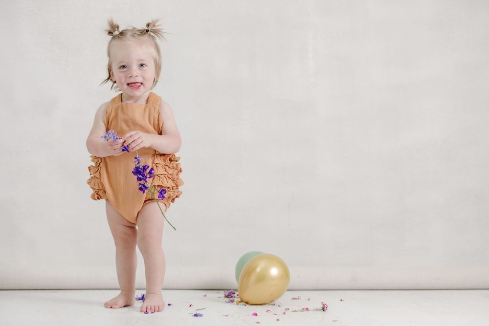 Small child in mustard yellow outfit standing next to a gold balloon with purple party decorations scattered on floor.