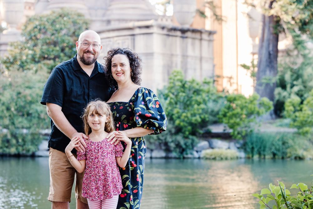 A family stands together smiling at the Palace of Fine Arts in San Francisco with a serene lagoon in the background.