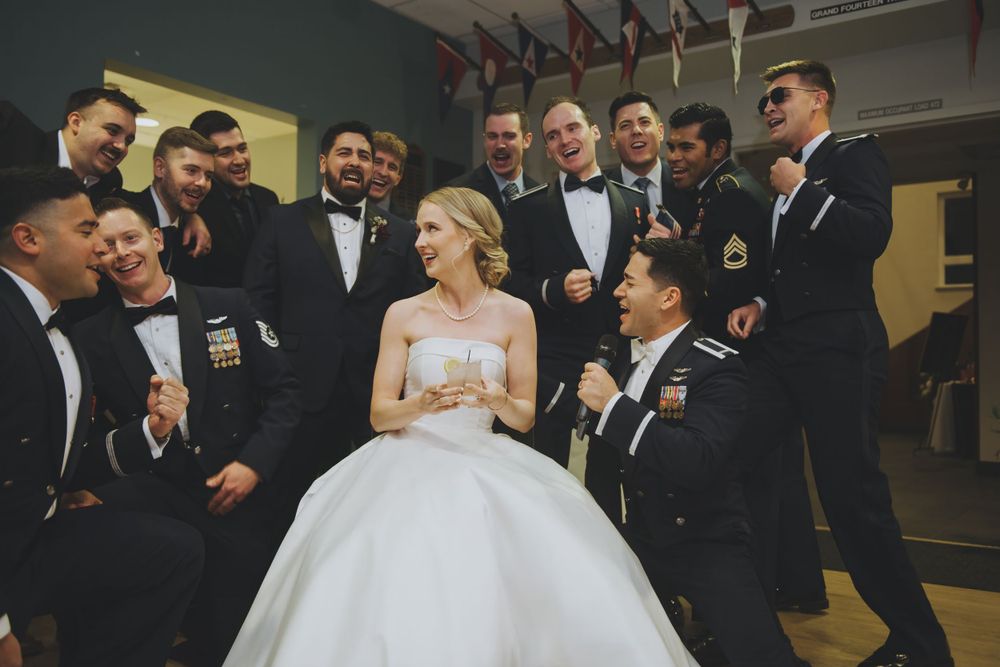 A bride in an elegant white ball gown poses with a group of groomsmen in black tuxedos at an indoor venue.