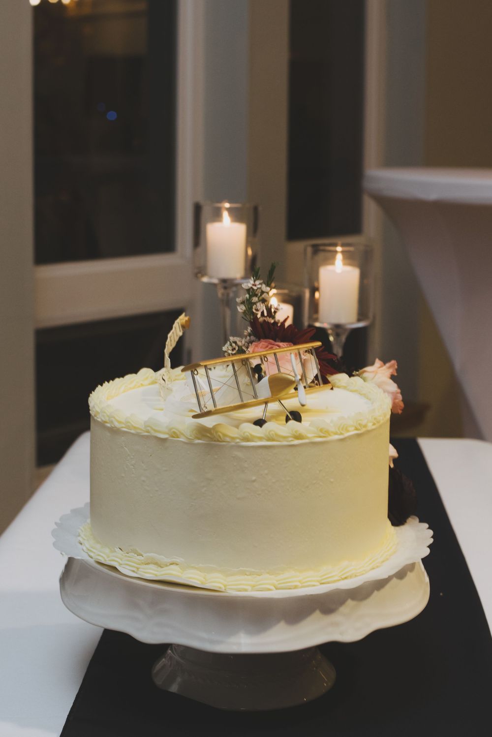White buttercream cake decorated with miniature airplane figures on a pedestal stand next to glowing candles.