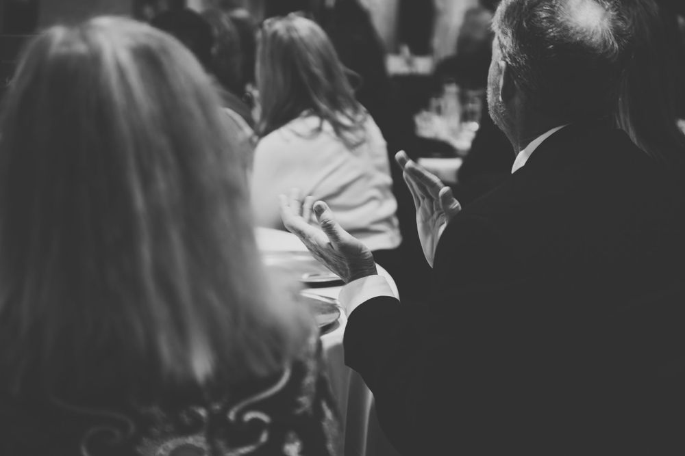 Black and white photograph of people seated in an audience, focused on the backs of their heads and shoulders.