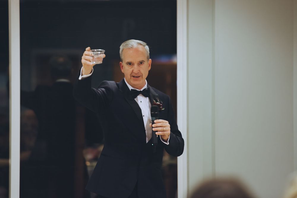The father-of-the-bride in black tuxedo raises a glass to toast the couple during a wedding reception.