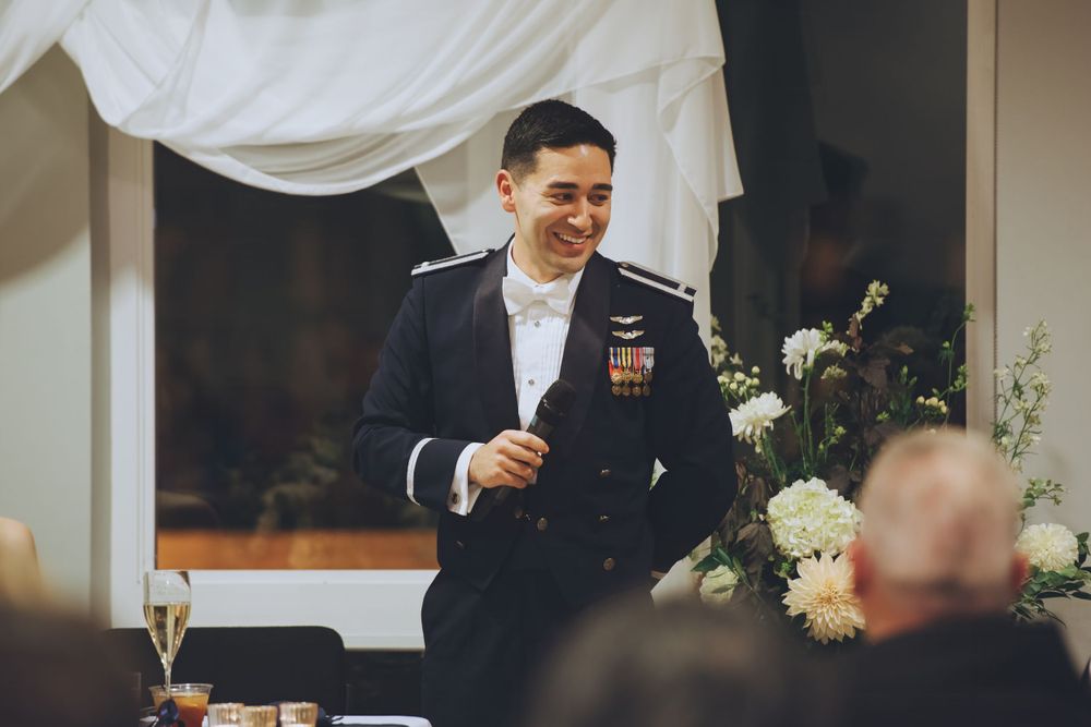 Groom in military dress uniform smiles while giving a speech at wedding reception with champagne glasses and floral decor.
