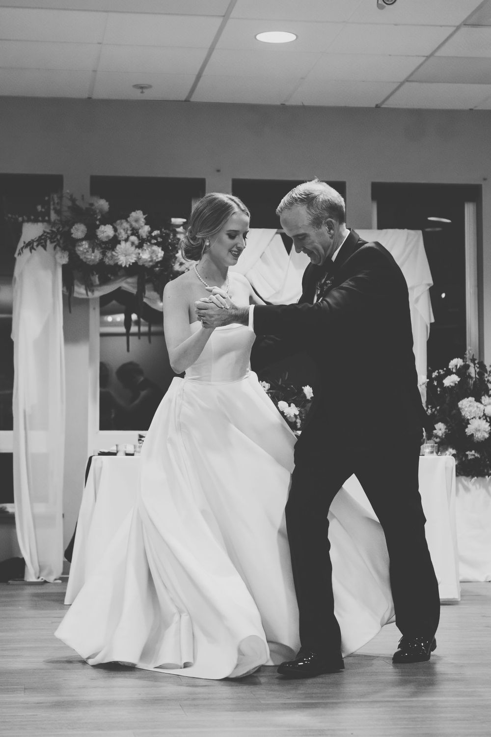 A bride in a flowing white gown dances with her father in a dark suit during a wedding reception.