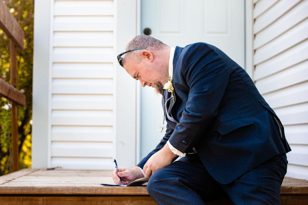 Person in navy suit writing on wooden porch steps against white building exterior.