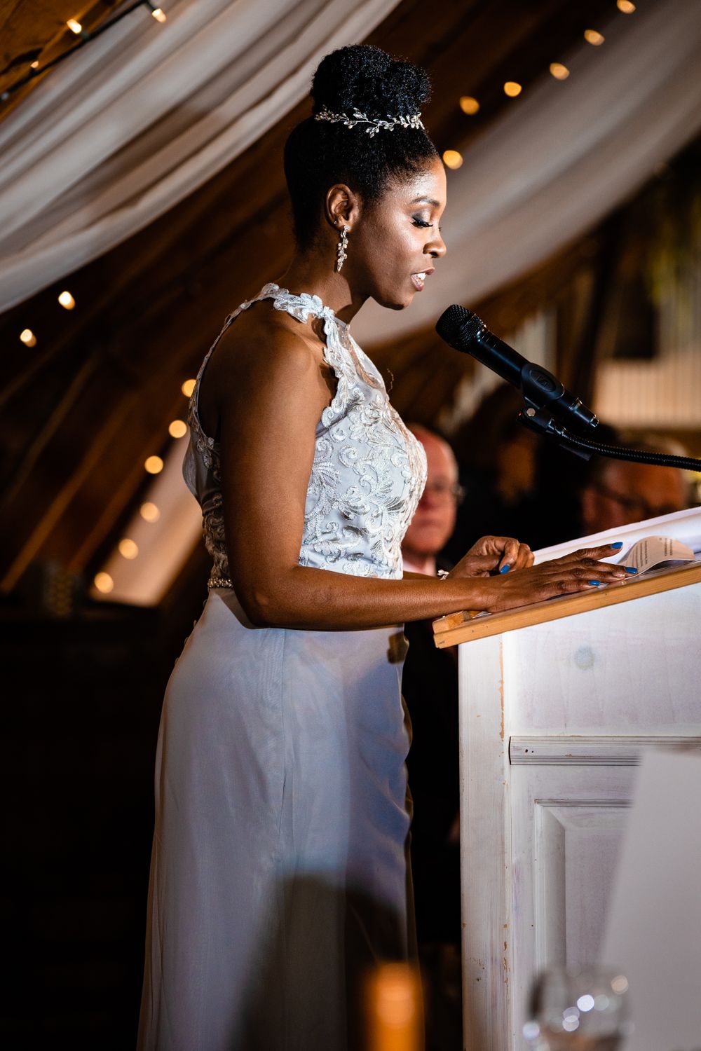Person in white lace wedding dress giving speech at elegant reception podium.