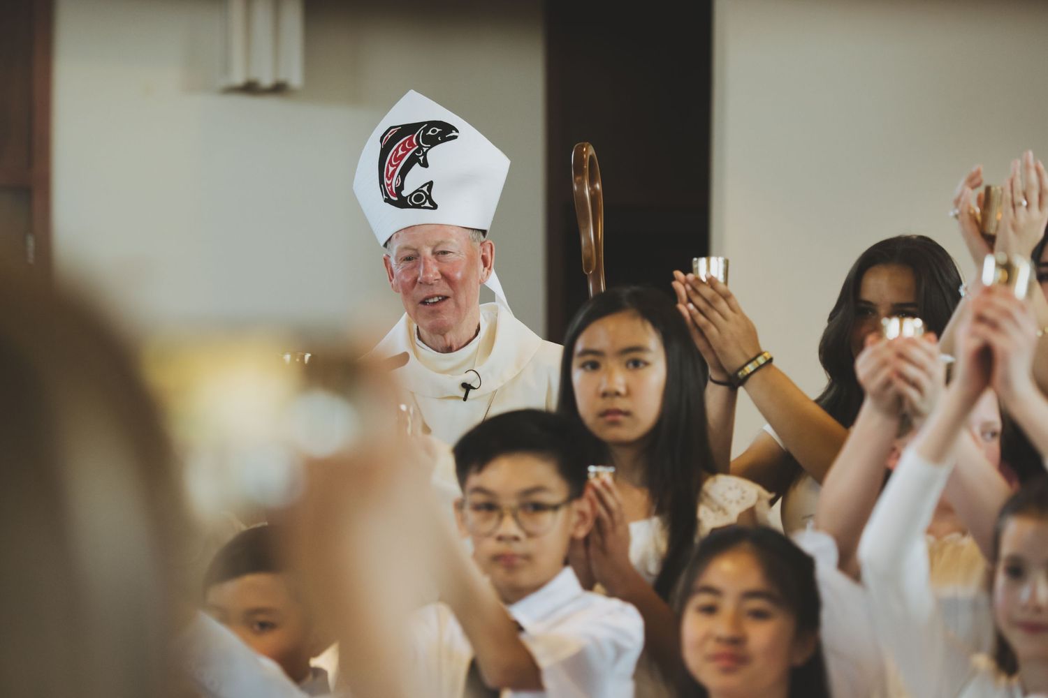 Students raise their hands in a church gathering with a bishop wearing a white mitre hat and clerical attire.